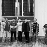 Seven men standing in a row outside a corrugated metal wall, posing for a group photo (likely a band). They wear casual clothes and jeans, with a large vent behind them.