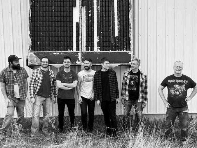 Seven men standing in a row outside a corrugated metal wall, posing for a group photo (likely a band). They wear casual clothes and jeans, with a large vent behind them.