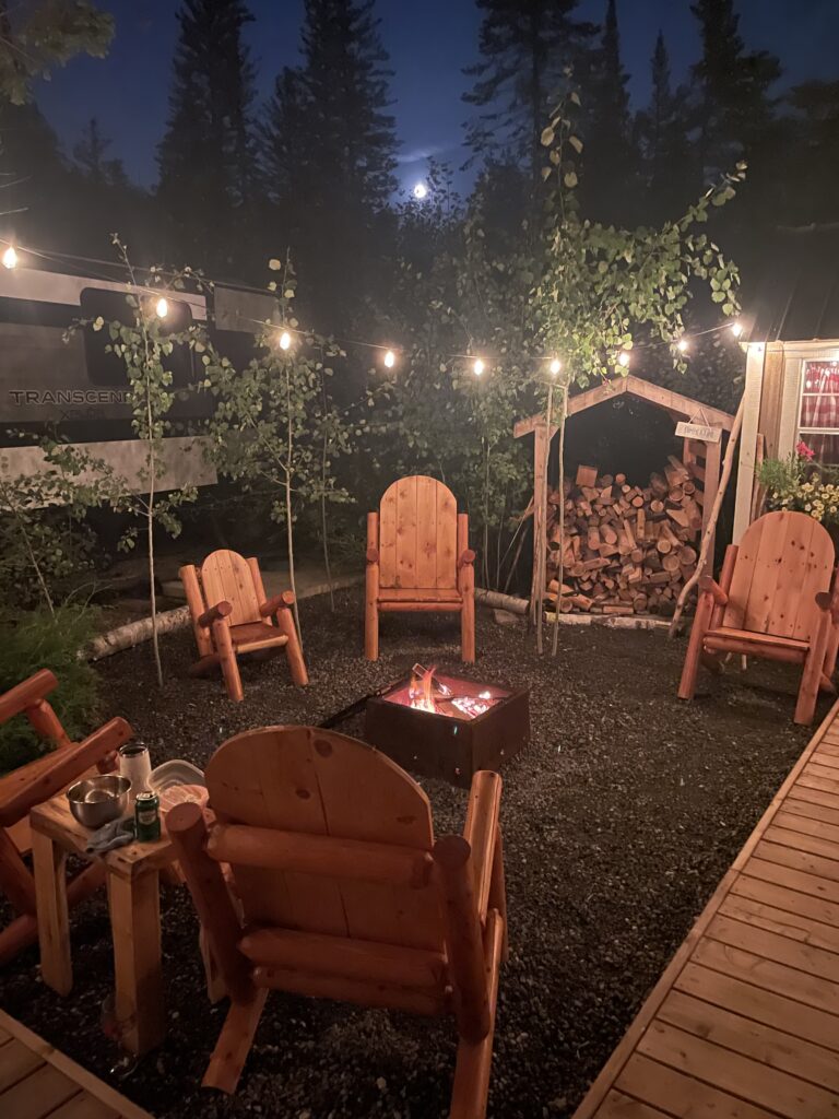 Night backyard scene with a glowing fire pit surrounded by wooden Adirondack chairs and string lights, logs stacked nearby.