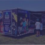 Outdoor event with a blue promotional trailer and people lining up near a purple inflatable obstacle course in a park urban area above grass under a clear sky.