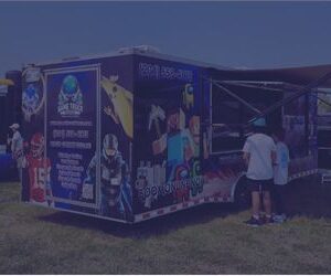Outdoor event with a blue promotional trailer and people lining up near a purple inflatable obstacle course in a park urban area above grass under a clear sky.