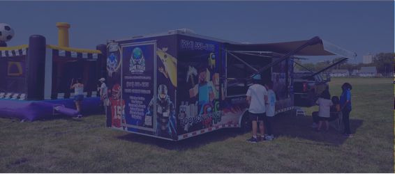 Outdoor event with a blue promotional trailer and people lining up near a purple inflatable obstacle course in a park urban area above grass under a clear sky.